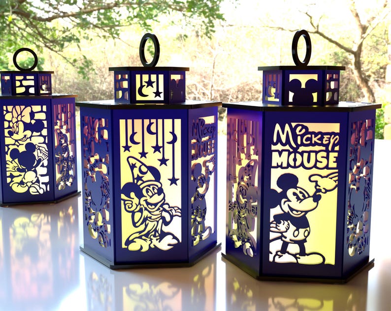 May include: Three blue decorative lanterns with cut-out designs featuring Mickey Mouse and other Disney characters. Each lantern has a black handle and a yellow interior, creating a warm glow. The lanterns are placed on a white surface.