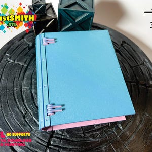 May include: A light blue notebook with a pink spine and three metal clasps. The notebook is sitting on a black surface with a 3D printed logo in the top right corner.