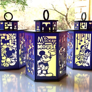 May include: Three blue lanterns with Mickey Mouse and friends designs. Each lantern has a black top with a circular handle. The sides feature cut-out images of Mickey, Minnie, and other characters. The lanterns are lit from within, creating a warm glow.