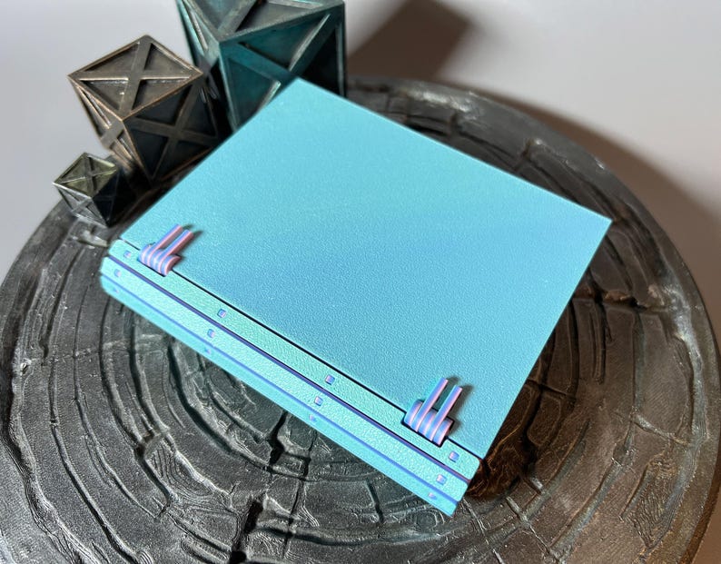 May include: A teal blue notebook with a textured cover and a unique binding system. The notebook has a subtle iridescent sheen and is closed.