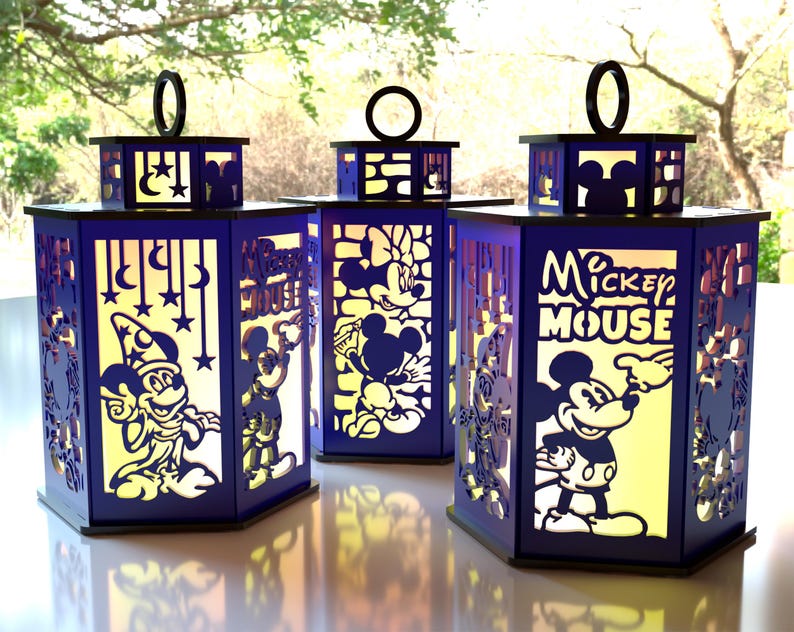 May include: Three navy blue Disney-themed lanterns with cut-out designs featuring Mickey Mouse, Minnie Mouse, and other characters. Each lantern has a black handle and a yellow glow from within, creating a whimsical effect.
