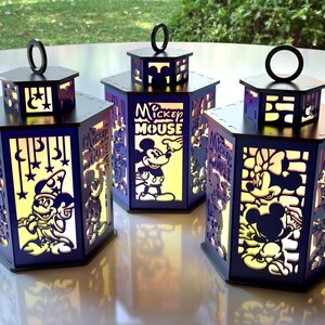 May include: Three illuminated, hexagonal lanterns with black tops and bases. The lanterns feature cut-out designs of Mickey Mouse, Minnie Mouse, and other Disney characters. The sides are a dark blue with yellow-toned light shining through the cutouts.