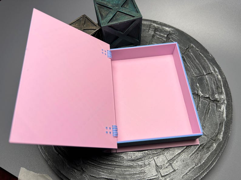 May include: A pink box with a blue and white trim. The box is open and the inside is empty. The box is sitting on a gray surface.