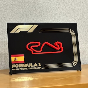 Puede incluir: Una placa negra con un logotipo de Fórmula 1 dorado y un contorno rojo del circuito de Barcelona-Cataluña. La placa también presenta la bandera española y el texto "FORMULA 1 GRAN PREMIO DE ESPAÑA".
