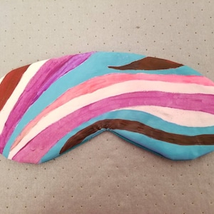 Puede incluir: Una colorida máscara para dormir con un diseño pintado. La máscara presenta un patrón de rayas curvas en tonos de azul, rosa, blanco y marrón. Tiene una banda elástica blanca para asegurar alrededor de la cabeza. La máscara está diseñada para cubrir los ojos.