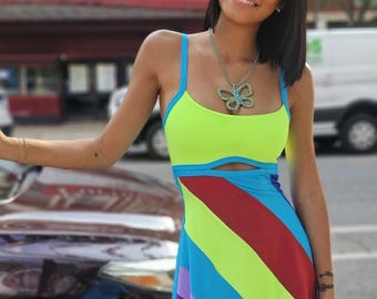 May include: A woman wearing a bright green, blue, purple, and red striped dress with a butterfly necklace. The dress has a cut-out detail at the waist.