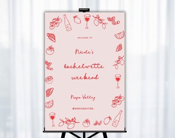 May include: A pink welcome sign with a red border and red illustrations of wine glasses, grapes, lemons, and tomatoes. The text on the sign reads "Welcome to Nicole's bachelorette weekend Napa Valley #MRSHASHTAG".