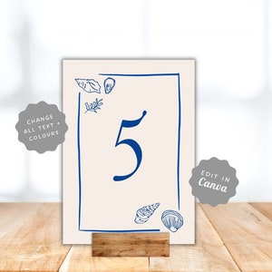 May include: A printable table number card with a blue border and a white background. The number 5 is written in blue inside the border. There are blue seashells and a blue leaf design on the card. The text "Edit in Canva" is on a grey circle in the bottom right corner.