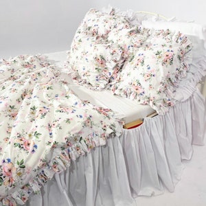 Könnte beinhalten: Weißes Bett mit einem weißen, gerüschten Bettrock und einer floral gemusterten Bettdecke und Kissen. Die Bettdecke und die Kissen haben einen weißen Hintergrund mit rosa, blauen und grünen Blumen.