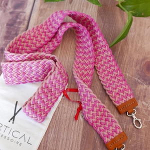 Peut inclure: Une sangle tressée rose et beige avec des fermoirs argentés et des accents de cuir marron. La sangle est faite d'un matériau épais et tressé et est conçue pour un sac. Un sac blanc avec du texte noir est également visible.
