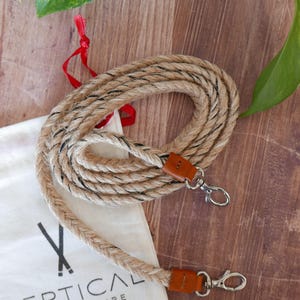 Peut inclure: Une sangle en corde enroulée avec des accents noirs et des extrémités en cuir, avec des fermoirs argentés. La sangle est sur une surface en bois, à côté d'un sac blanc avec le texte "VERTICAL L'ACCESSOIRE MADE IN FRANCE". Un ruban rouge est également visible.