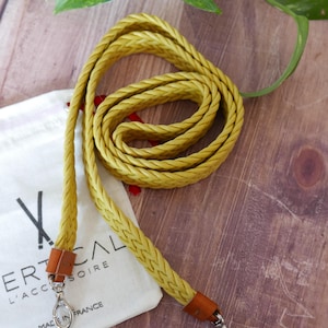 Peut inclure: Une lanière en cuir jaune tressé avec des accents en cuir marron et des fermoirs argentés. La lanière est enroulée sur une surface en bois, avec un sac blanc portant le texte "VERTICAL L'ACCESSOIRE MADE IN FRANCE".