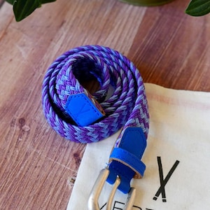 Peut inclure: Une ceinture tressée présentant un motif tissé de fils violets et bleu clair. La ceinture comprend un embout en similicuir bleu vif et un passant en similicuir bleu assorti près d'une boucle en métal argenté à ardillon unique.