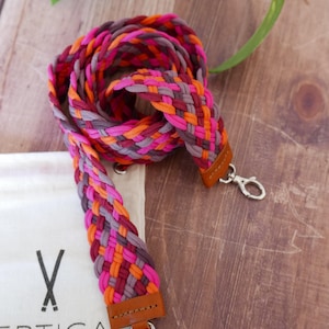 May include: Braided strap in shades of pink, orange, and gray with brown leather ends and silver clasps. The strap is lying on a wooden surface next to a white bag with the text "VERTICAL D'ACCESSOIRE" and "MADE IN FRANCE".