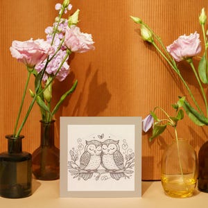 May include: A black and white illustration of two owls cuddling on a branch with flowers and leaves. The illustration is printed on a white card with a grey border and is displayed on a table with pink flowers in vases.