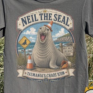 May include: A charcoal gray short-sleeve t-shirt featuring a graphic of a seal wearing an orange traffic cone on its head. The text reads NEIL THE SEAL, KEEP BACK I DO WHAT I WANT, and TASMANIA'S CHAOS ICON, with a yellow seal warning sign.