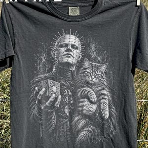 Può includere: Una t-shirt grigio antracite con una stampa grafica bianca di un personaggio con spilli in testa che tiene in mano un cubo rompicapo, con un gatto tigrato nell'altro braccio. La parola GRAPHITE è stampata in bianco in alto a sinistra.