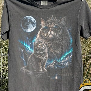 May include: A charcoal gray t-shirt featuring a graphic print of a large cat wearing round glasses and a crown, positioned above a smaller cat sitting on a rock. The scene includes a full moon, a mountain landscape, and blue aurora borealis lights.