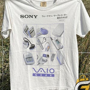 Puede incluir: Una camiseta blanca de manga corta con una cuadrícula de nueve ilustraciones de dispositivos electrónicos Sony antiguos. Sobre la cuadrícula, el texto dice SONY y caracteres japoneses. Debajo, el texto en morado dice VAIO GEAR.