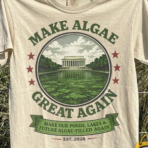 May include: A cream-coloured short-sleeve t-shirt featuring a circular graphic of the Lincoln Memorial surrounded by green algae. The text reads "MAKE ALGAE GREAT AGAIN" and "MAKE OUR PONDS, LAKES & FUTURE ALGAE-FILLED AGAIN" with "EST. 2026" below.
