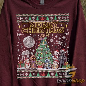 Op de afbeelding: Een bordeauxrode sweater met ronde hals en een grafische print met de tekst MERRY CHRISTMAS. Het ontwerp bevat een kerstboom in Star Wars-thema, de Death Star, een AT-AT walker en diverse personages waaronder Darth Vader en R2-D2.