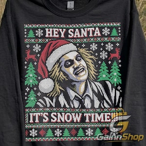 Puede incluir: Una sudadera negra de manga larga con un estampado gráfico de un personaje de pelo revuelto con un gorro de Papá Noel. El diseño incluye el texto HEY SANTA e IT'S SNOW TIME, rodeado de patrones de pinos verdes, renos rojos y copos de nieve blancos.