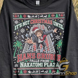 May include: A black long-sleeve sweatshirt featuring a graphic print of a man in a suit and Santa hat falling from a skyscraper. The text reads IT'S NOT CHRISTMAS UNTIL HANS GRUBER FALLS FROM NAKATOMI PLAZA, surrounded by festive patterns of trees and reindeer.