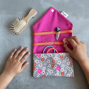 May include: A pink and grey fabric pouch with a floral pattern and the word "Hago" printed on the front. The pouch is open and contains hair ties and clips.