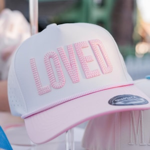 以下が含まれることがあります： ピンク色の「LOVED」の文字が刺繍された白とピンクの野球帽。帽子はカーブしたつばと白いロープのディテールが特徴です。帽子は透明なスタンドに展示されています。