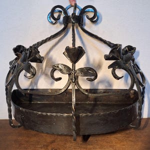 Può includere: Un cestino da parete in ferro battuto scuro con dettagli floreali e a volute. Il cestino ha un design curvo e aperto per contenere oggetti, con rose e foglie decorative. La lavorazione del metallo include un supporto centrale.