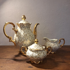 May include: A vintage tea set featuring a teapot, sugar bowl with lid, and creamer. The white porcelain pieces are adorned with a gold floral pattern and gold trim. The set is displayed on a wooden surface.