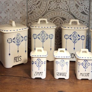Può includere: Un set di barattoli da cucina in ceramica bianca e una bottiglia con motivi decorativi blu e bianchi. Il set include contenitori con le etichette "ESSIG", "REIS", "ZIMMT", "GEWURZ", "MUSCAT", "ZUCKER" e "KAFFEE".