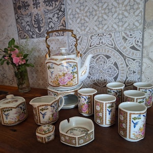 May include: A white porcelain teapot with floral and bird designs, a matching sugar bowl, creamer, and six teacups. The teapot has a gold handle and spout. The teacups have floral and bird designs. The set is on a wooden surface.