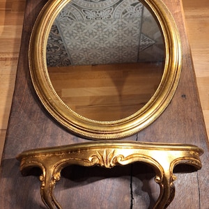 May include: An oval gold mirror with a decorative gold shelf beneath it. The mirror has a detailed frame and reflects a patterned ceiling. The shelf features ornate detailing and curved legs, mounted on a dark wooden surface.