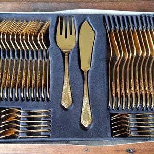 Könnte beinhalten: Ein komplettes goldfarbenes Besteckset in einer blau samtgefütterten Box. Das Set enthält Gabeln, Messer und Löffel, alle mit dekorativen Griffen. Die Anordnung deutet auf eine formelle Essumgebung hin, ideal für besondere Anlässe.
