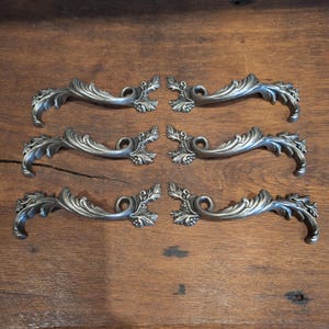 Pode incluir: Seis puxadores de gaveta prateados antigos com um design decorativo de folhas e videiras. Os puxadores estão dispostos em pares, mostrando suas alças curvas e ornamentadas e seus detalhes. Eles estão sobre uma superfície de madeira quente.