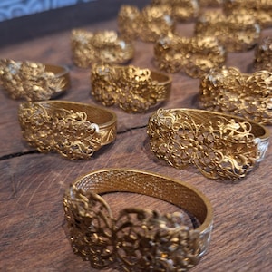 May include: Several gold-coloured metal bracelets with elaborate, openwork floral patterns. The bracelets are displayed on a dark wooden surface, highlighting their detailed craftsmanship and vintage style. Each bracelet is a unique piece.