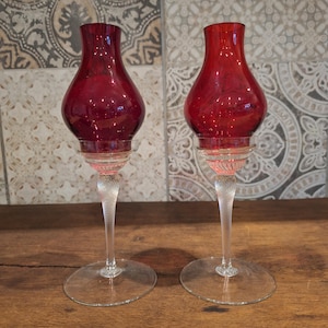 Könnte beinhalten: Zwei rubinrote Glas-Kerzenhalter. Jeder hat einen bauchigen Aufsatz, einen klaren Glasstiel mit gedrehtem Design und einen kreisförmigen Sockel. Die Kerzenhalter werden auf einer Holzoberfläche vor einem gemusterten Fliesen-Hintergrund präsentiert.