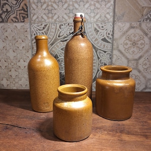 May include: Four antique stoneware bottles and jars in varying sizes. The vessels are a warm, earthy brown color with a speckled texture. One bottle has a swing top closure. The items are arranged on a wooden surface.