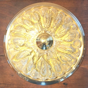 Könnte beinhalten: Eine runde, goldfarbene Deckenleuchte mit floralem Design. Die Leuchte hat eine zentrale, reflektierende Kuppel und ist von einem Ring aus strukturierten, blütenblattartigen Glaselementen umgeben. Das Licht ist auf einer Holzoberfläche montiert.
