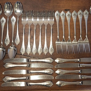 Può includere: Una collezione di posate antiche, tra cui cucchiai, forchette e coltelli, disposte su una superficie di legno. Le posate presentano dettagli ornati e una finitura lucida. Un set completo per la cena.