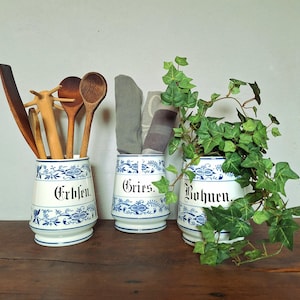 Könnte beinhalten: Drei weiße Keramikbehälter mit blauen Blumenmustern und schwarzer deutscher Beschriftung Erbsen, Gries und Bohnen. Die Behälter enthalten hölzerne Küchenutensilien, gefaltete graue Stoffe und eine grüne Efeupflanze mit herabhängenden Blättern.