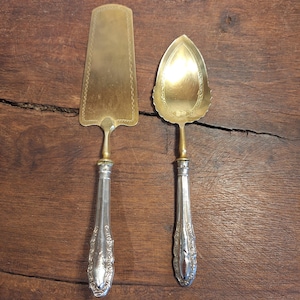 Può includere: Due utensili antichi da portata: una paletta per torte e una paletta per crostate. La paletta per torte ha una lama rettangolare dorata con un bordo decorativo. La paletta per crostate ha una lama dorata a forma di foglia. Entrambi hanno manici argentati ornati.