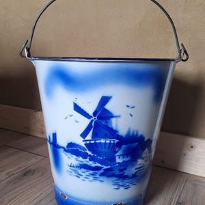 Op de afbeelding: Een wit en blauw geëmailleerde emmer met een windmolenscène. De emmer heeft een metalen handvat en een blauwe rand en bodem. De windmolenscène is blauw geschilderd en toont een windmolen, huizen en water.