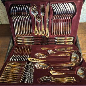 May include: A complete gold and silver-toned flatware set, neatly arranged in a burgundy velvet-lined case. The set includes forks, knives, spoons, and serving utensils. The handles feature an ornate gold design, adding a touch of elegance to the dining set.