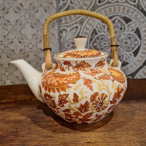 Pode incluir: Uma chaleira branca com um padrão floral em tons de laranja, amarelo e marrom. A chaleira tem uma alça de bambu e uma tampa.