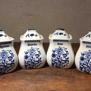Può includere: Quattro barattoli di spezie in ceramica bianca con coperchi, ciascuno etichettato con un nome di spezia diverso in un carattere a script. I barattoli presentano un design floreale blu e sono disposti su una superficie di legno.