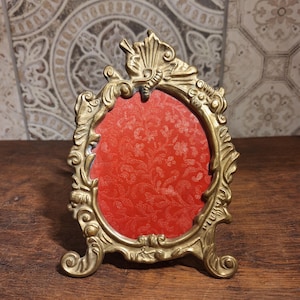 Puede incluir: Marco de fotos dorado antiguo y ornamentado, con una abertura ovalada. El marco tiene elaborados adornos y una parte superior decorativa. El interior presenta un fondo rojo estampado. El marco está diseñado para colocarse de pie sobre una superficie plana.