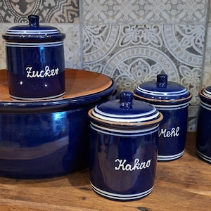 Könnte beinhalten: Fünf blaue Keramikdosen mit weißen Deckeln und weißer Beschriftung. Die Dosen sind mit "Zucker", "Kakao", "Mehl" und "Salz" beschriftet.