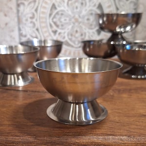 Puede incluir: Un conjunto de cuencos de postre de acero inoxidable con base de pedestal. Los cuencos tienen una superficie brillante y reflectante y están colocados sobre una mesa de madera. El fondo presenta una pared de azulejos estampados.